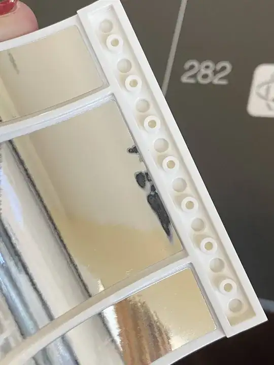 Fingers holding a lego piece with mirrored stickers applied on the inner side of the LEGO. There are three stickers and the center, largest sticker has slight bubbles, which are noticeable due to the warped reflection in the sticker