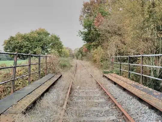 Picture looking down an abandoned railway and railway bridge on a grey autumn day with some yellow leaves on the ground and the trees on the edges of the image are green and yellow and orange.