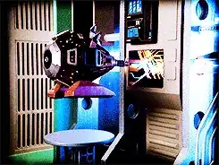 An excomp, looking like a mostly black faceted spheroid with blinking lights, silver details and a pair of large red pyramidal feet, hovers in the air in front of a vertically-mounted console. A cylindrical device on its "front" is projecting a blue beam that is triggering patterns of lines in the display.