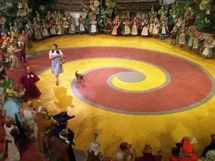 Movie still showing the red and yellow brick roads in a spiral pattern entwined together, Dorothy and Toto set off down the yellow brick road with a crowd encircling them