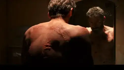 A screen capture from the aforementioned trailer from the upcoming movie "Logan" (2016), showing the titular character, Logan (also known as Wolverine), standing at a dimly lit mirror, his back visibly and heavily scarred.