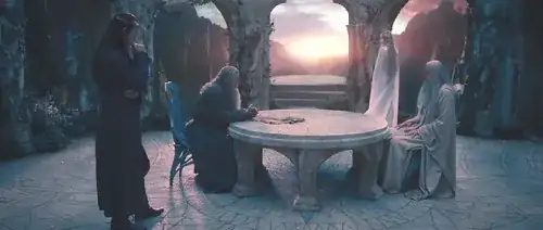 Gandalf (seated left, leaning forward) and Saruman (seated right) face one another across a circular table.  Elrond stands watching them from Gandalf's right, slightly behind, while Galadriel stands in the background, facing out a window at the setting sun.