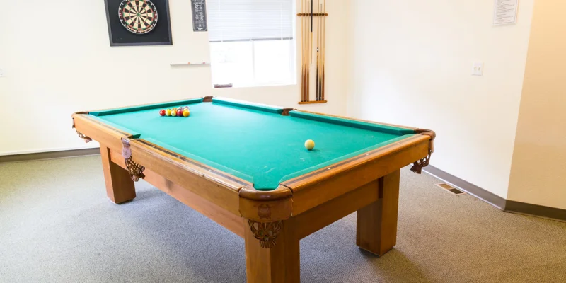 A pool table in a recreational room with a dartboard and cue sticks.