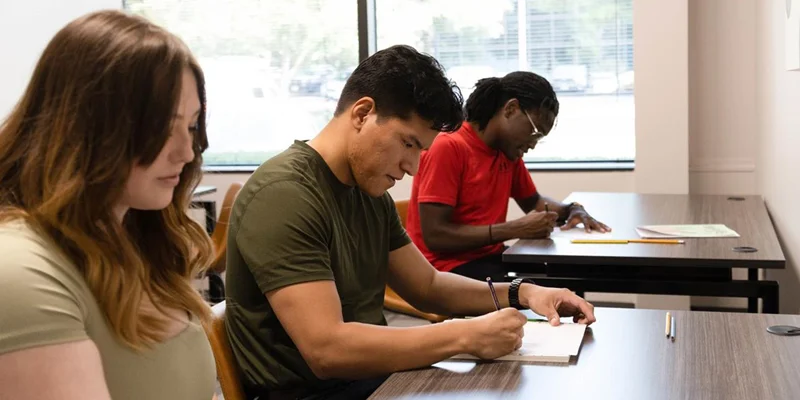 Three people writing at wooden desks in a bright room.