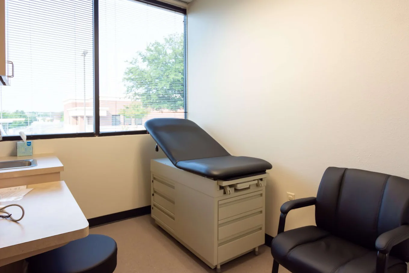 A medical exam room with a black exam table and chairs near a window
