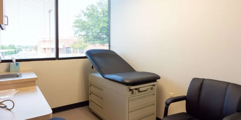 A medical exam room with a black exam table and chairs near a window