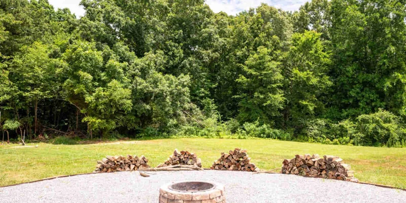 Outdoor fire pit with gravel and firewood, surrounded by trees.