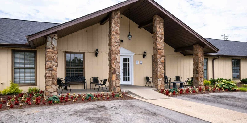 Exterior of a rehab facility with stone columns, a sitting area with black chairs, and landscaping