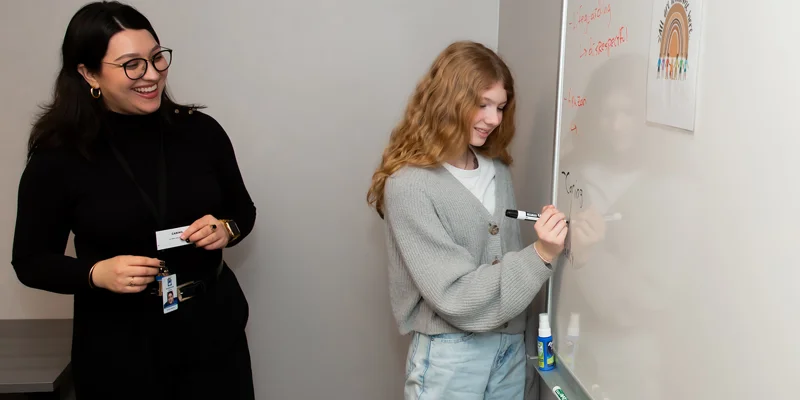 A young woman writes on a whiteboard while another person observes.