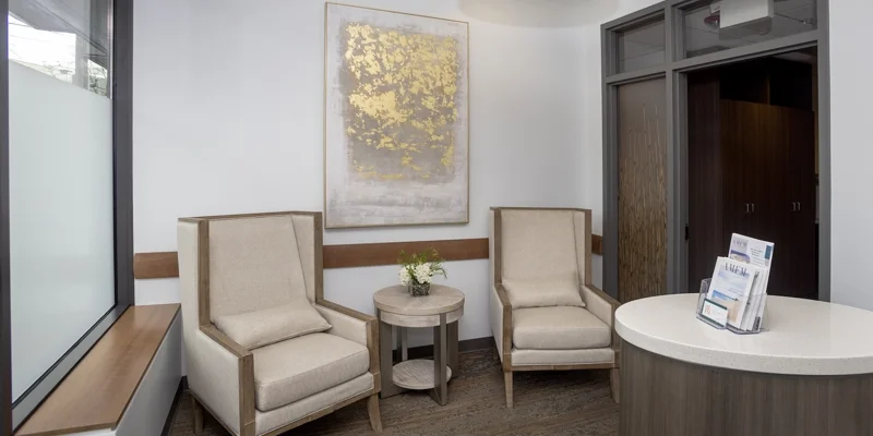 A waiting area with two beige chairs and a decorative gold painting