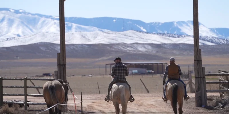 Two men on horseback ride away under a large wooden ranch archway.