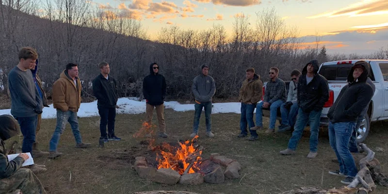 Group of men gathered around a campfire at dusk near snowy trees.