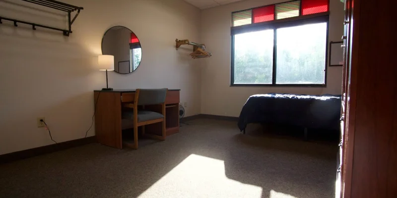 A bedroom with a desk, chair, mirror, and a single bed by the window