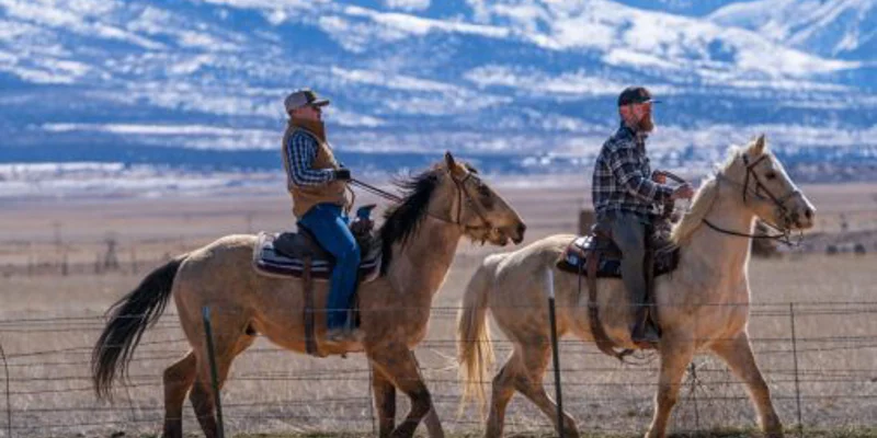 Two men riding horses in a wide, open field with snowy mountains behind.