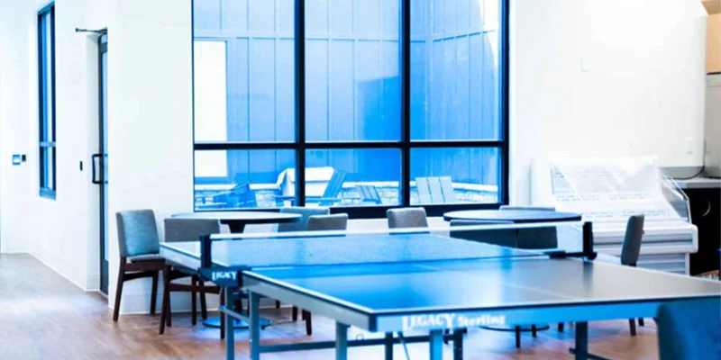 A rec room with a ping-pong table, chairs, and large windows.