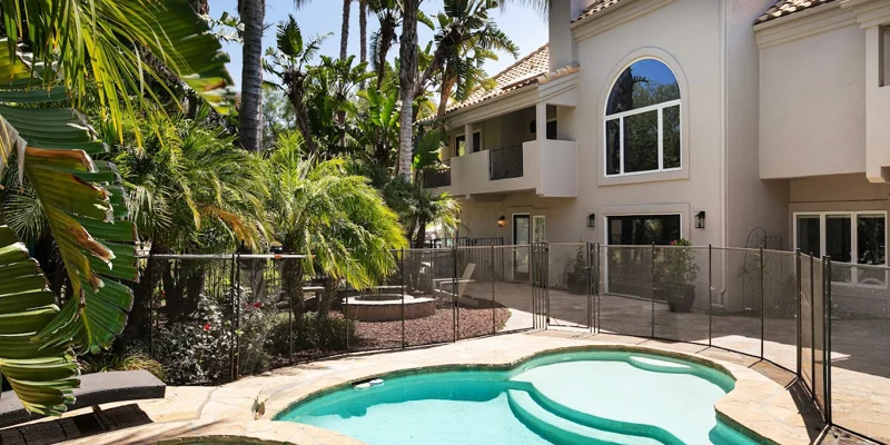 Outdoor pool area with palm trees and a fenced patio beside a villa.