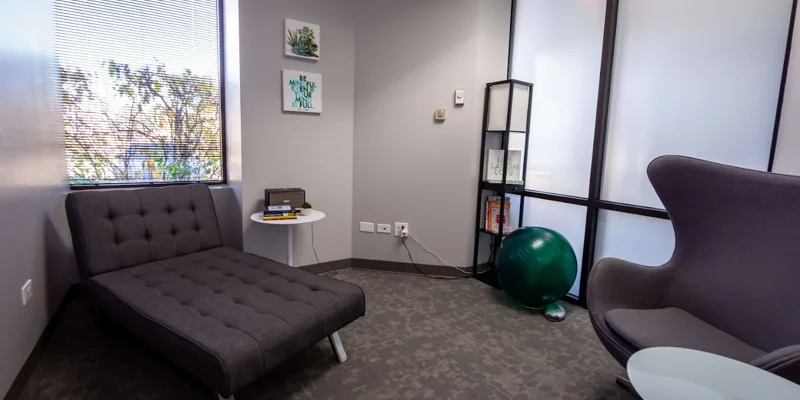 Therapy room with a gray chaise, modern chair,  and large windows.