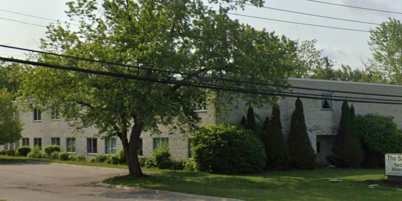 A light-colored building partially hidden by large trees and bushes, with a sign in front.