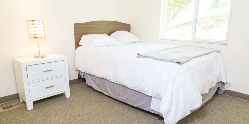 A bedroom with a white bed, nightstand, and window with natural light.