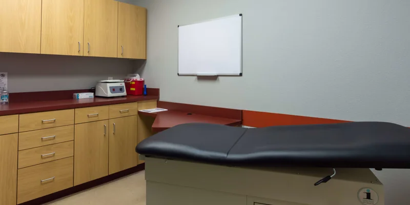 A medical exam room with a black examination table and wooden cabinets.