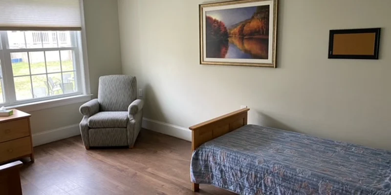 A simple bedroom with a twin bed, chair, and framed artwork.