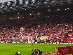 A stand which is full of people standing to support their team. There are a number of flags, scarfs, and banners in the crowd.