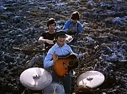 The Beatles performing music in a field. In the foreground, the drums are played by Starr (only the top of his head is visible). Beyond him, the other three stand in a column with their guitars. In the rear, Harrison, head down, strikes a chord. In the front, Lennon smiles and gives a little wave toward camera, holding his pick. Between them, McCartney is jocularly about to choke Lennon.