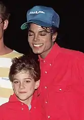 A smiling Jackson wears a blue baseball cap and a red shirt. On his left, a young boy smiles. He is dressed in a red shirt, too.