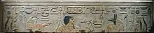 Light gray stone surface with carved and painted images of two woman, a falcon-headed god, a black-haired man with a long goatee, a jackal-headed god, and Egyptian hieroglyphs inscribed along the top