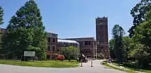 A red brick building is seen, with grass, trees, and construction equipment in the foreground.  The building's tower and main entrance are to the right, with glass roofs and windows stretching to the left.  Architecturally, the tower echoes the shape of the tower in the original building.