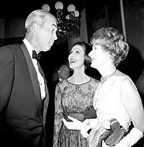 Monochrome photo of two women and a man dressed in formal attire - the two women (standing right) are smiling up at the man (facing opposite), who looks slightly amused.