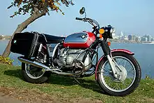 Red BMW R75/5 motorcycle fitted with panniers, parked on grass by a lake with a city in the background
