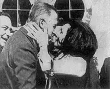 Profile of a woman resting her hand on the neck of a man facing her and rubbing her nose against his, while two other men look on.