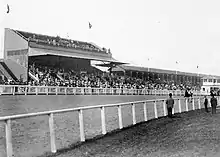 Black and white photo of horses running around a dirt track with a grandstand full of spectators in the background