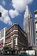 In the foreground: the abandoned Yoyogi Kaikan&nbsp;[ja] building that appears in the film. The demolition of the building was completed in January 2020 and it no longer exists. Note that the rooftop shrine in this film is fictional. Back: the NTT DoCoMo Yoyogi Building (Docomo Tower), which frequently appears in Shinkai's films.