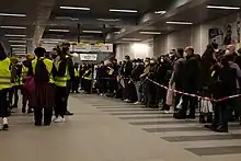 Viele Menschen hinter einer Absperrung, auf der anderen Seite Helfer mit Westen und Megaphonen