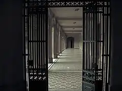 Galería techada vista a través de una puerta de rejas. A la izquierda se ven unas columnas. En el fondo se ve una puerta de madera.