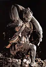 Fūjin. Three-quarter view of a statue. His left leg is bend as if climbing stairs and he is carrying a long bag-shaped object which goes from one shoulder to the other around the back of his head. Black and white photograph.