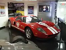 Vehículo rojo deportivo con dos rayas blancas e identificado con el número 1, en exhibición dentro de una sala con otros automóviles.