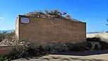 Se observa una construcción en forma rectangular con vegetación en la parte superior. En la construcción hay un cartel que indica la calle. Alrededor hay más vegetación. No se aprecia que haya sido usada.