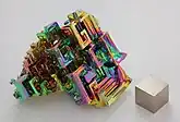 Bismuto en forma cristalina, con una capa de oxidación muy fina, y un cubo de bismuto de 1&nbsp;cm3