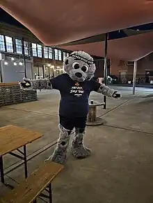 Una mascota con la forma de una foca antropomórfica de pie con las manos a los lados y vistiendo una camisa azul oscuro que dice Never say die ("Nunca digas morir")