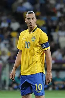 A photograph of a man with dark hair wearing a yellow football shirt, blue shorts and a dark blue captains armband on his arm, the man is looking away from the camera.