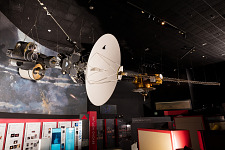 Antenna on white disc with an aluminum arm that supports multiple instruments, including cameras and spectrometers. A long boom extends on the right over an exhibit.