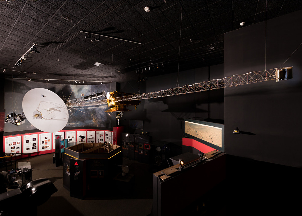 Antenna on white disc with an aluminum arm on the left that supports multiple instruments. A long magnetometer boom extends on the right over the exhibit space below.