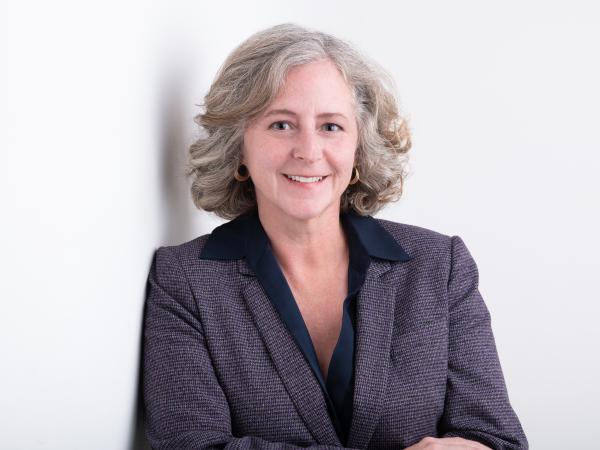 Elizabeth Babcock from the waist up against a white background. She is leaning against a wall with her arms crossed in front of herself.  