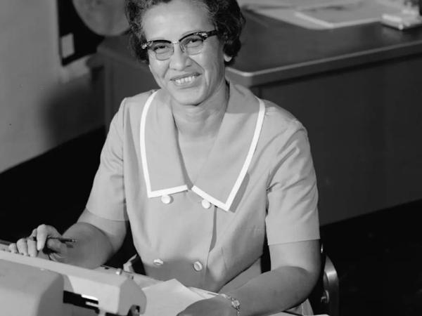 A black and white photo of Katherine Johnson sitting at a desk in front of a typewriter and open notebook.