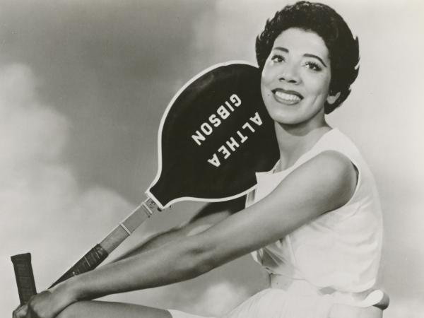 In this black-and-white photograph, Althea Gibson poses on a stool with two tennis rackets. Both the rackets have dark colors printed with Althea Gibson in white letters.