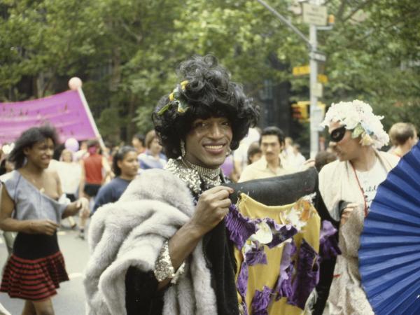 Marsha P. Johnson looks directly at the camera, smiling, and holding a black quilt with gold, purple, and white embellishments, while she herself wears black apparel with silver sequin collar and cuffs, with rhinestone earrings and necklaces.