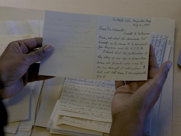 Two hands holding a card with handwritten information. A stack of other handwritten papers is visible on the table below. 